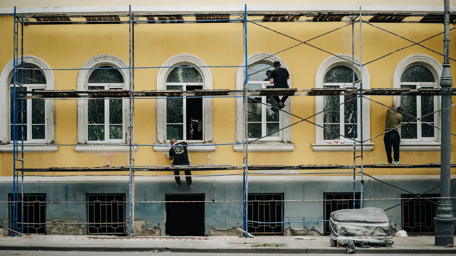 Εργάτες σε σκαλωσιά επισκευάζουν την πρόσοψη κίτρινου κτιρίου.