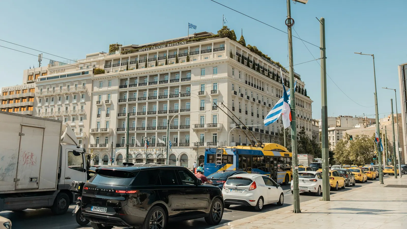 Δρόμος της Αθήνας με κίνηση, μεγάλο κτίριο και ελληνική σημαία.
